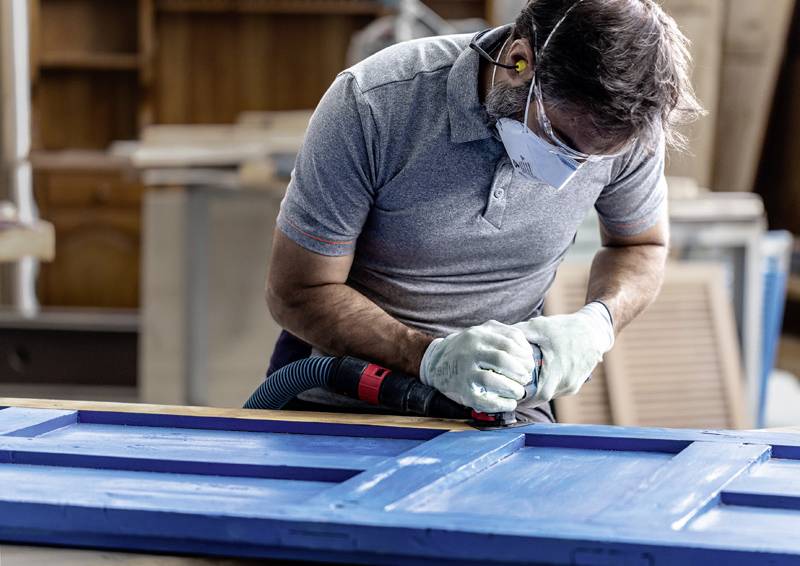 Eine Person, die eine Maske und Handschuhe trägt, benutzt einen elektrischen Elektroschleiffer an einer blauen Holztür in einer Werkstattsituation.