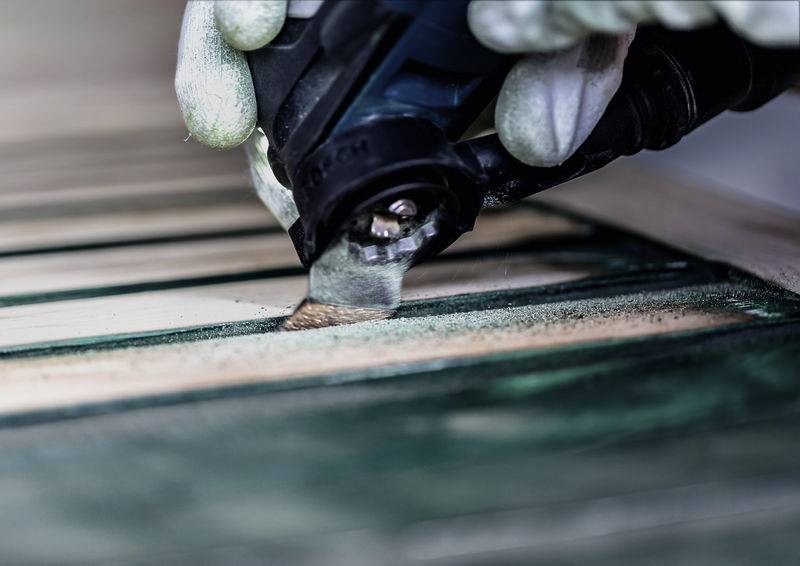 Eine Hand in Handschuhen benutzt ein Elektrowerkzeug, um grüne Holzbretter zu schleifen, ihre Oberfläche zu glätten und Farbe oder Lack zu entfernen.