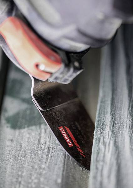 Eine Nahaufnahme einer Person, die eine Säge mit rotem Griff verwendet, um ein Holzstück durchzusägen, mit sichtbarem Sägemehl auf der Oberfläche.