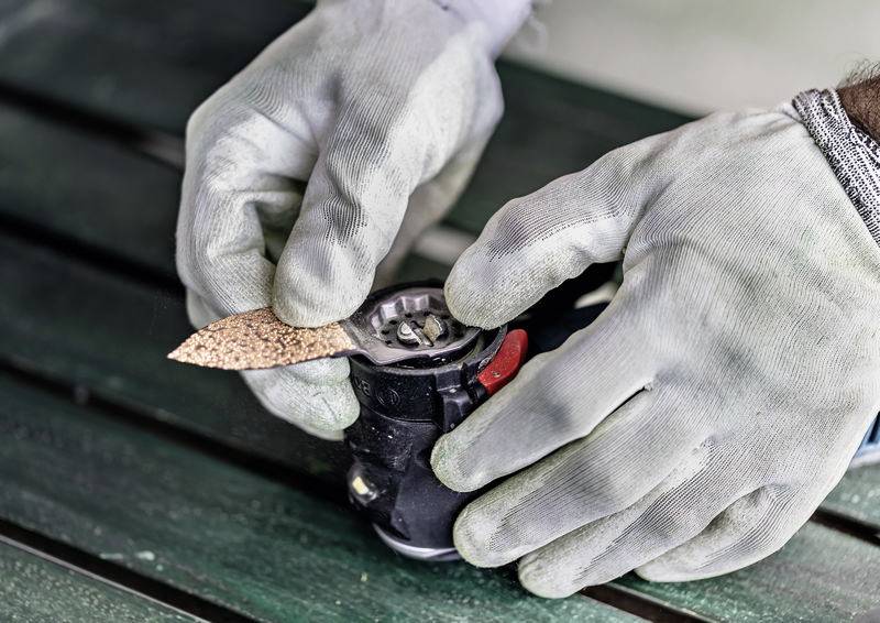 Hände in Schutzhandschuhen, die Schleifpapier an einem elektrischen Schleifen anpassen, mit Fokus auf den Mechanismus des Werkzeugs und die körnige Textur.