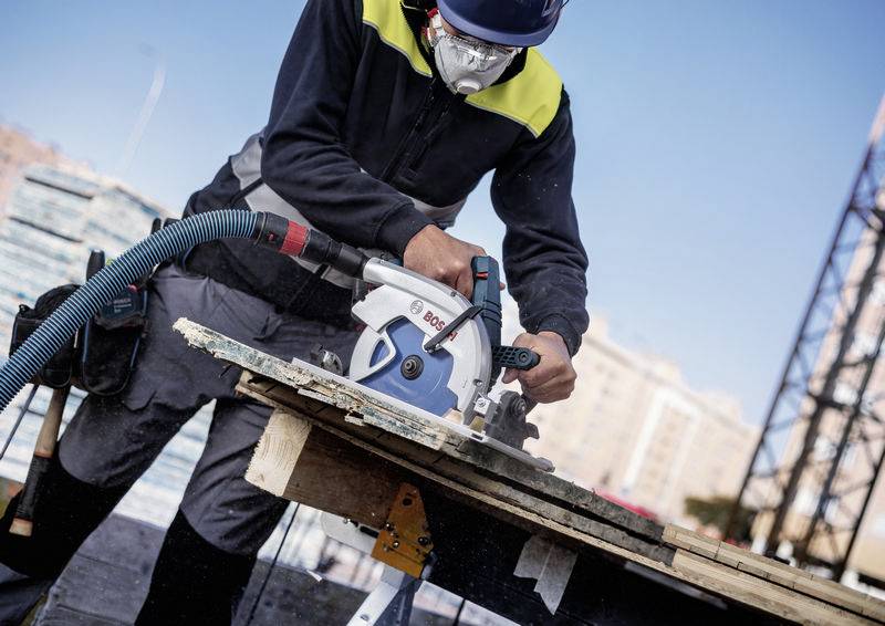 Ein Arbeiter benutzt eine Kreissäge, um Holz auf einer Baustelle zu schneiden, wobei er eine Schutzmaske, Handschuhe und eine Schutzbrille trägt.
