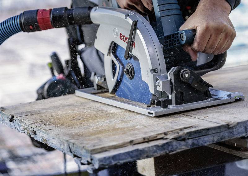 Eine Person benutzt eine Kreissäge, um ein Holzbrett durchzuschneiden, wobei Sägespäne umherwirbeln. Die Säge trägt die Marke „Bosch