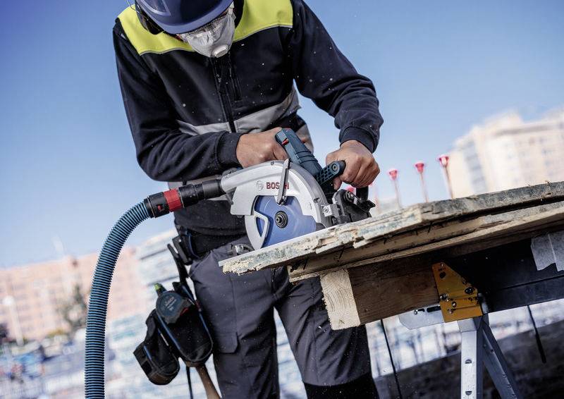 Eine Person, die mit einer Kreissäge Holz auf einer Baustelle schneidet, trägt Schutzausrüstung. Im Hintergrund sind städtische Gebäude zu sehen.