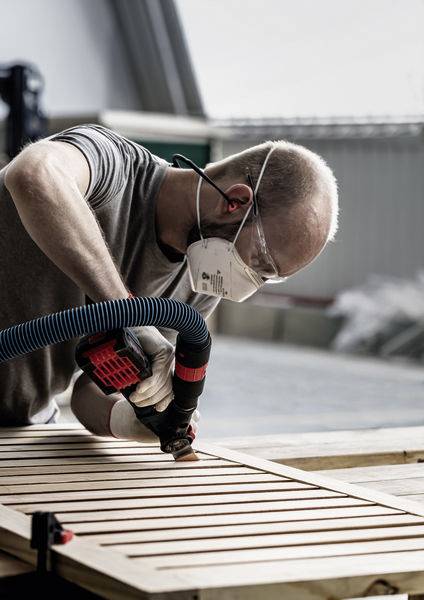 Eine Person mit Maske und Gehörschutz schleifen eine Holzoberfläche in einer Werkstatt, was auf eine Holzbearbeitungstätigkeit hindeutet.