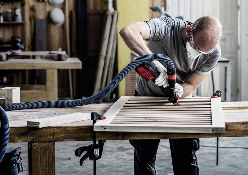 Eine Person, die Schutzausrüstung trägt, schleifen eine Holztür mit einem Elektrowerkzeug in einer Werkstatt und konzentriert sich auf die Aufgabe inmitten verschiedener Holzbearbeitungswerkzeuge.
