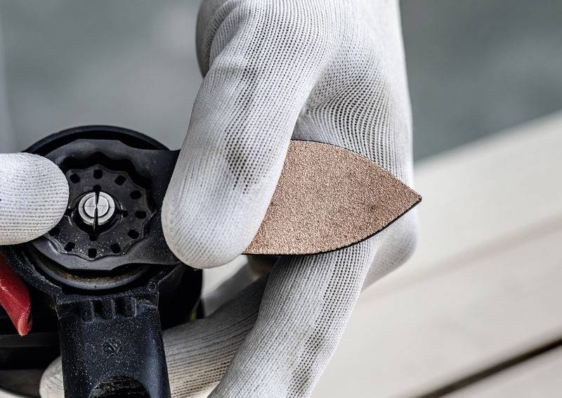 Eine behandschuhte Hand hält ein braunes blattförmiges Schleifpapierstück gegen ein schwarzes Schleifwerkzeug, was auf Wartungs- oder Reparaturarbeiten hindeutet.
