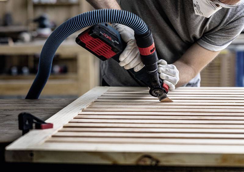 Eine Person, die Handschuhe und eine Maske trägt, benutzt eine elektrische Säge mit Staubsaugeransatz, um eine Holzleiste in einer Werkstattumgebung zu schneiden.
