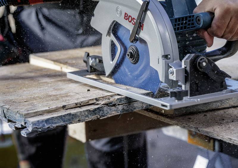 Eine Person schneidet ein Holzbrett mit einer Kreissäge. Das Brett zeigt sichtbare Abnutzung, und Sägespäne sind in der Luft. Die Säge trägt die Bezeichnung „BOSCH