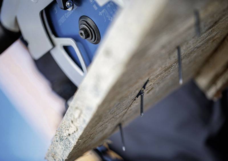 Eine Kreissäge schneidet durch ein Holzbrett, wobei Nägel aus dem Brett herausragen, was auf ein laufendes Bau- oder Zimmermannsvorhaben hindeutet.