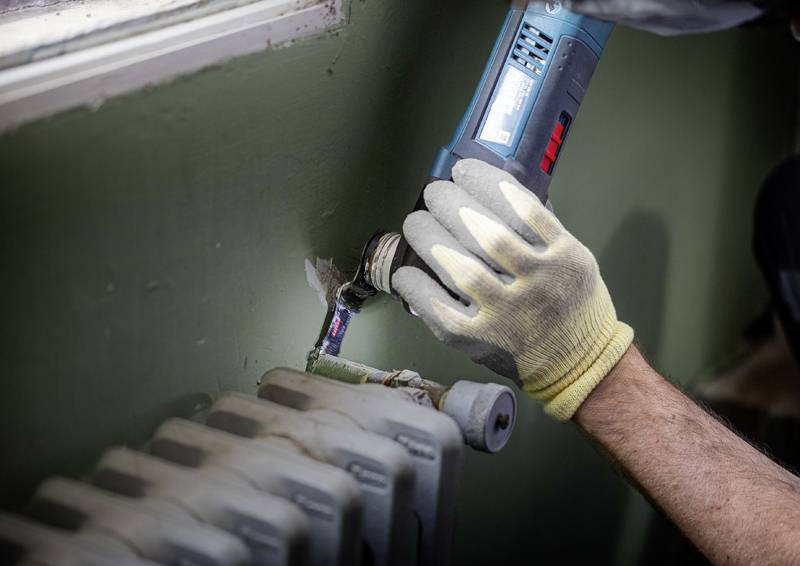 Eine Person mit Handschuhen benutzt ein blaues Elektrowerkzeug, um ein Rohr in der Nähe eines Heizkörpers in einem Raum zu reparieren, was auf Wartungsarbeiten hindeutet.