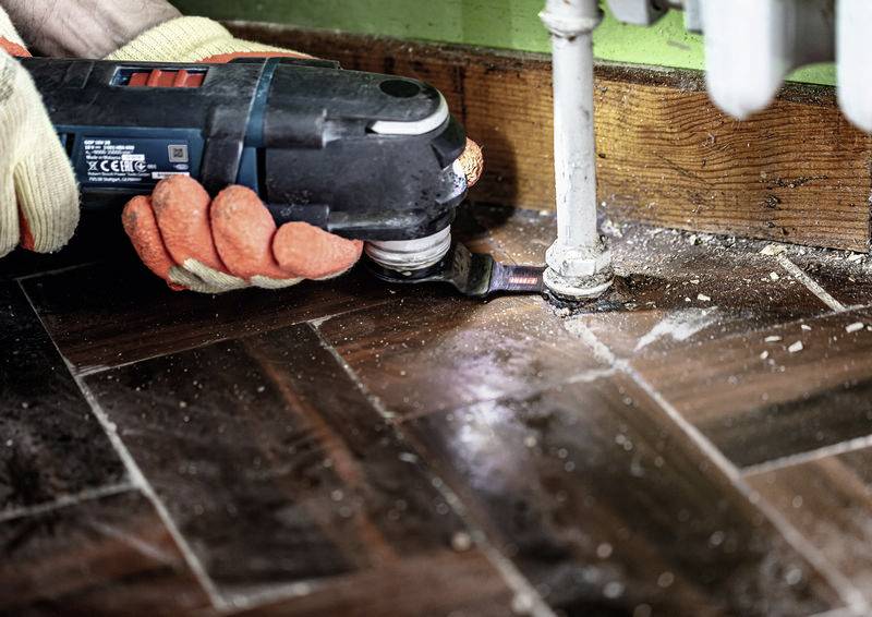 Eine Person mit Handschuhen benutzt ein Elektrowerkzeug, um an einem Rohr zu arbeiten, das auf einem Holzboden mit Fischgrätmuster montiert ist.
