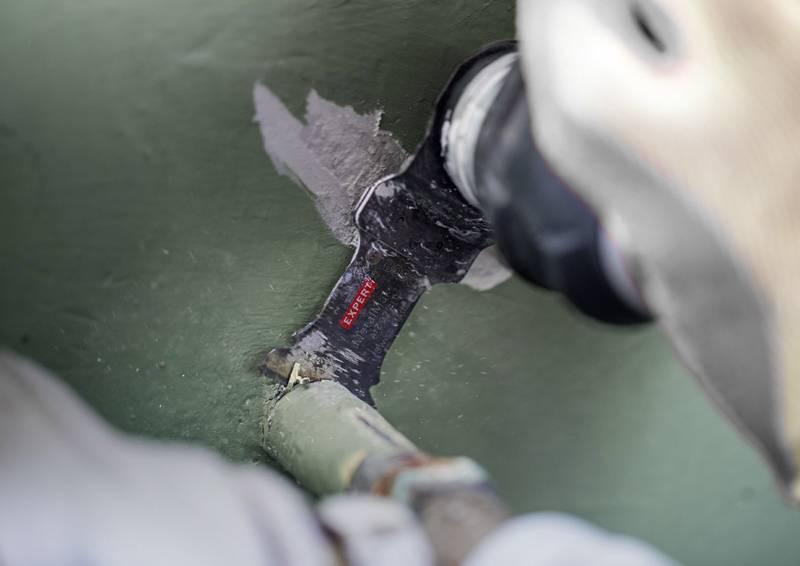 Eine Person mit Handschuhen benutzt ein Elektrowerkzeug, um alte Farbe von einem Rohr in einer Werkstatt zu entfernen.