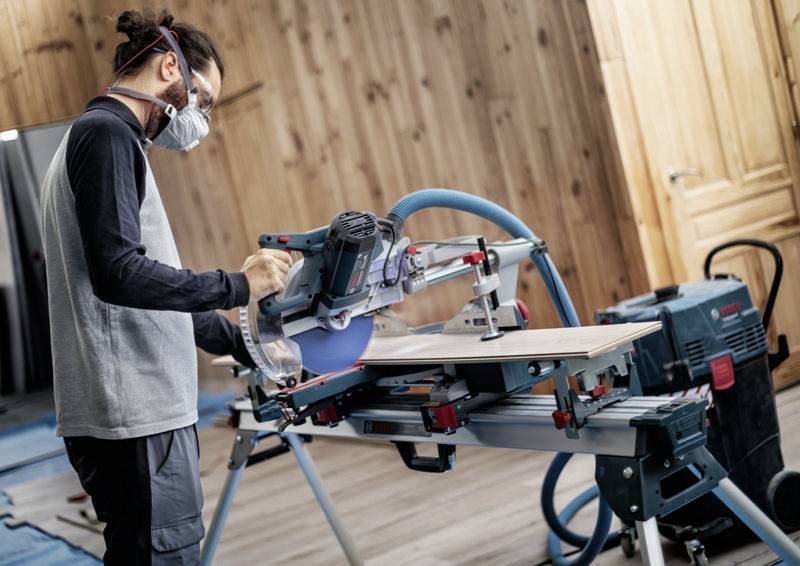 Eine Person mit einer Maske bedient eine Kreissäge in einem holzgetäfelten Raum und ist mit Holzbearbeitung beschäftigt.