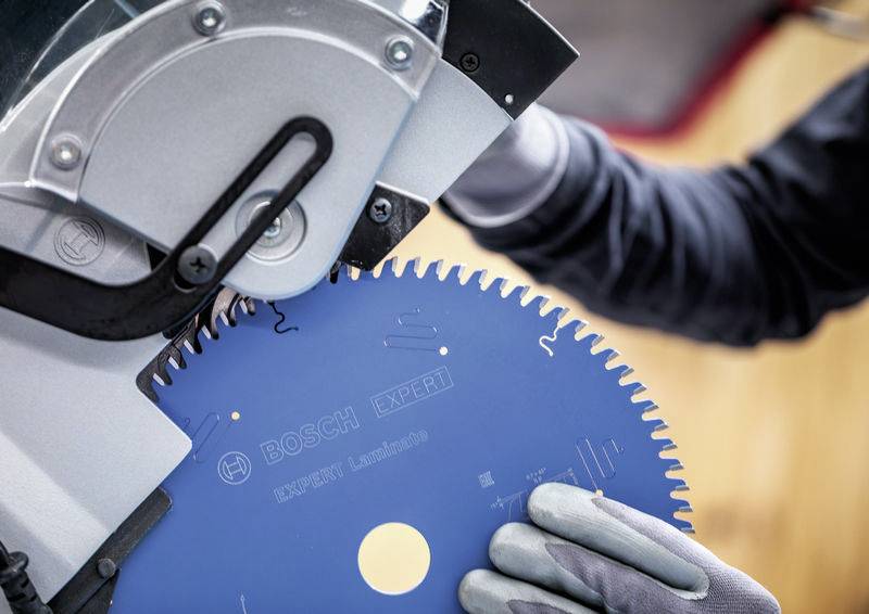 Eine Person mit Handschuhen setzt ein blaues Sägeblatt in eine Bosch-Schneidemaschine ein, möglicherweise in einer Werkstatt oder auf einer Baustelle.