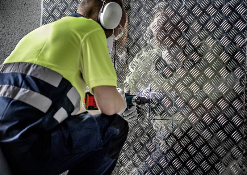 Eine Person trägt Schutzausrüstung und verwendet ein Elektrowerkzeug, um ein Quadrat aus einem strukturierten Metallblech zu schneiden, möglicherweise für Bau- oder Installationsarbeiten.