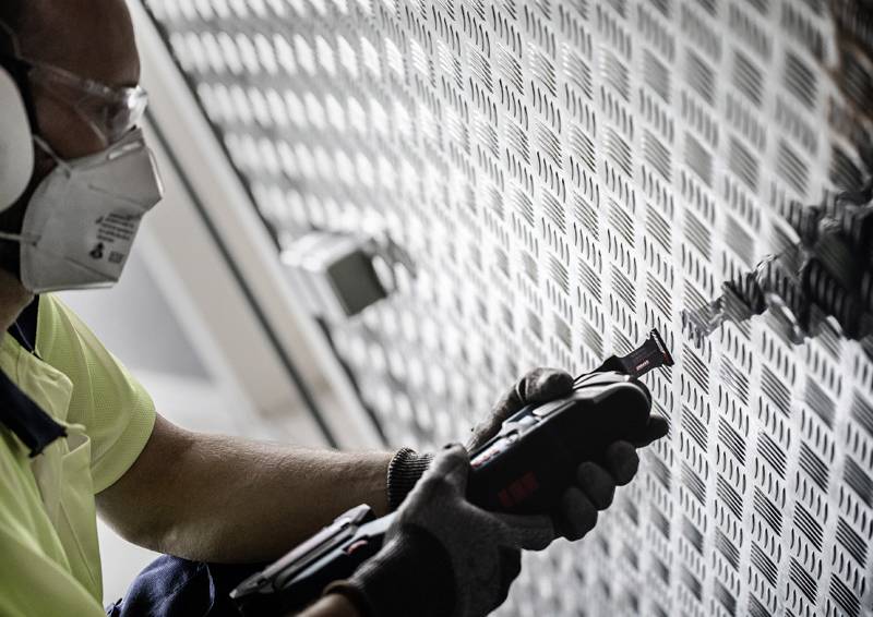 Arbeiter mit Maske und Handschuhen verwendet Elektrowerkzeug auf gemustertem Metallblech und konzentriert sich intensiv auf die präzise Schneid- oder Bohraufgabe.
