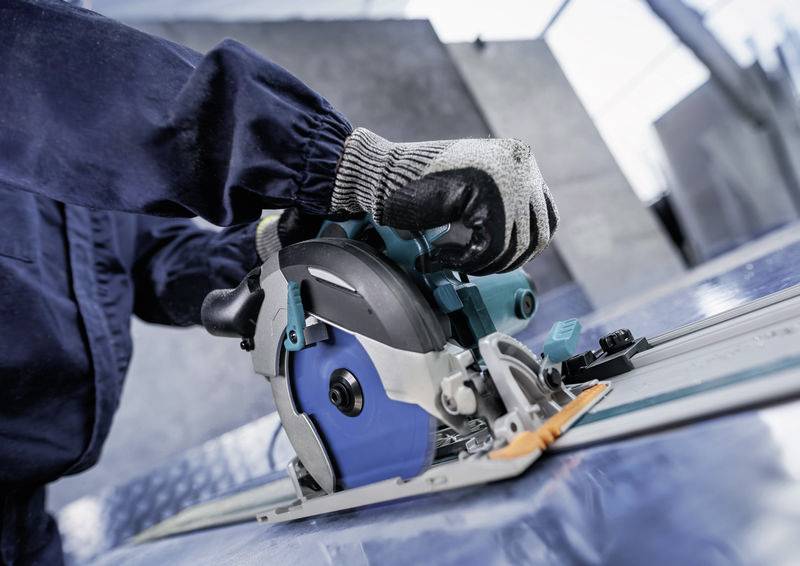 Eine Person mit Handschuhen bedient eine blau und silbern gefärbte Kreissäge und schneidet ein Metallblatt auf einer Werkbank in einer Industrieumgebung.