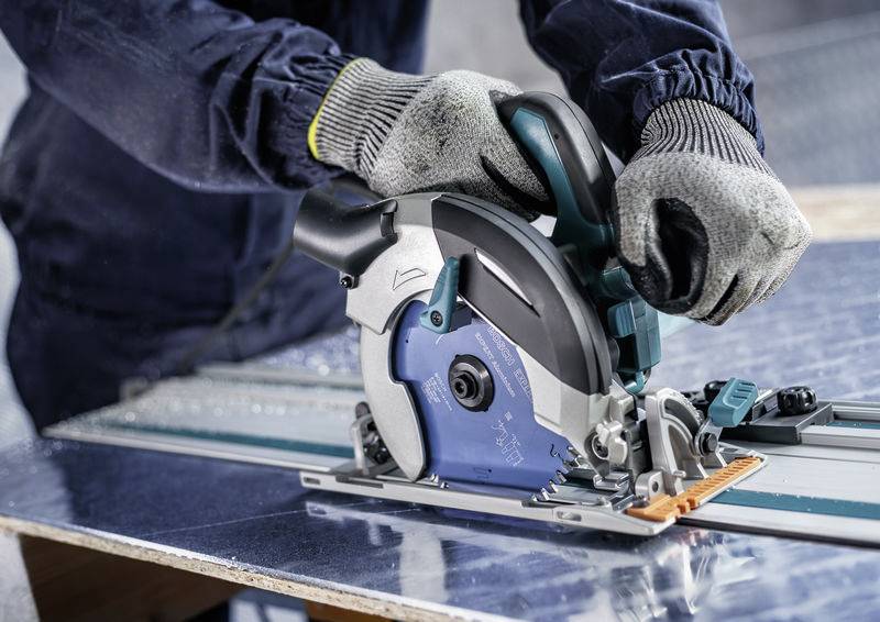Eine Person in Handschuhen bedient eine Kreissäge und schneidet ein Metallblech auf einer Werkbank. Sicherheitsausrüstung und Präzision sind deutlich erkennbar.