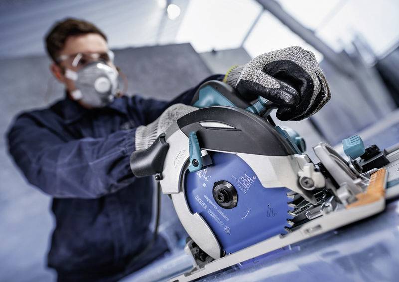 Eine Person in Schutzausrüstung bedient eine Kreissäge und schneidet Material auf einer Werkbank. Die Sicherheitsausrüstung umfasst Handschuhe und eine Maske.