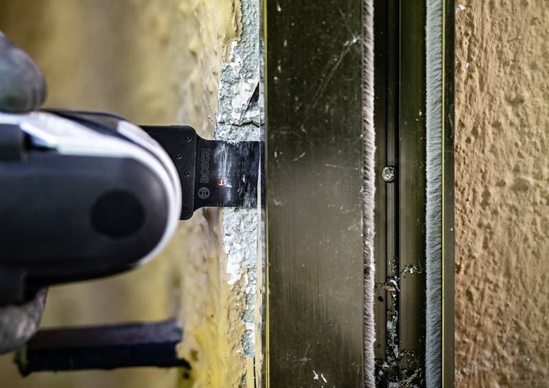 Ein Nahaufnahme-Foto zeigt ein Elektrowerkzeug, das in Gipskartonplatte neben einem Metallrahmen schneidet. Staub und Trümmer sind an der Schnittstelle sichtbar.