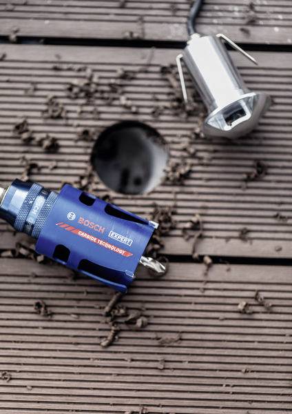 „Eine blaue Bosch-Bohraufsatz auf einer Holzoberfläche neben einem gebohrten Loch und einer Metallvorrichtung, mit Holzspänen ringsum verstreut.