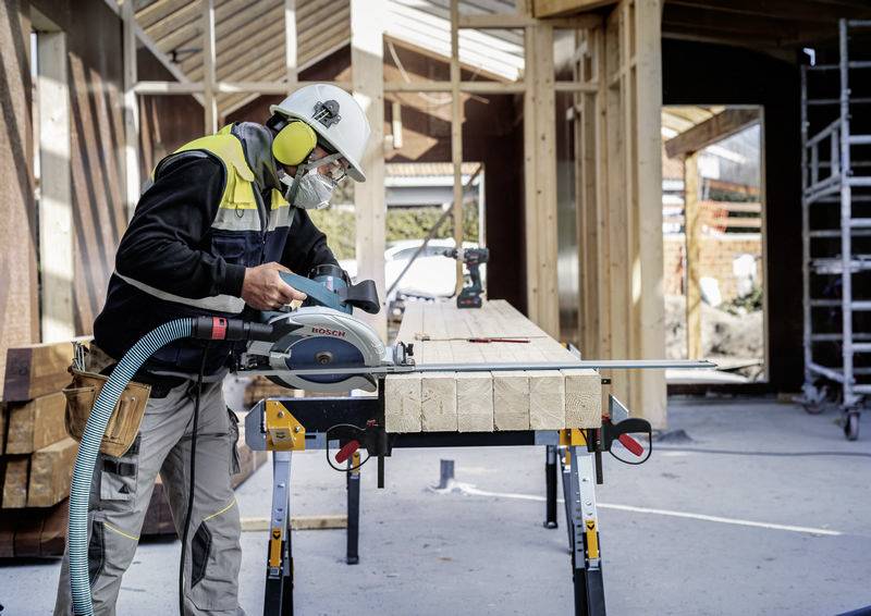 Arbeiter, der eine Kreissäge an hölzernen Balken auf einer Baustelle benutzt, trägt Sicherheitsausrüstung und ist von unfertigen Gebäuderahmen umgeben.