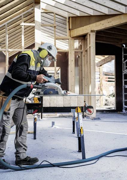 Ein Bauarbeiter in Schutzausrüstung schneidet Holz mit einer Kreissäge auf einer Baustelle, umgeben von teilweise errichteten Holzstrukturen.