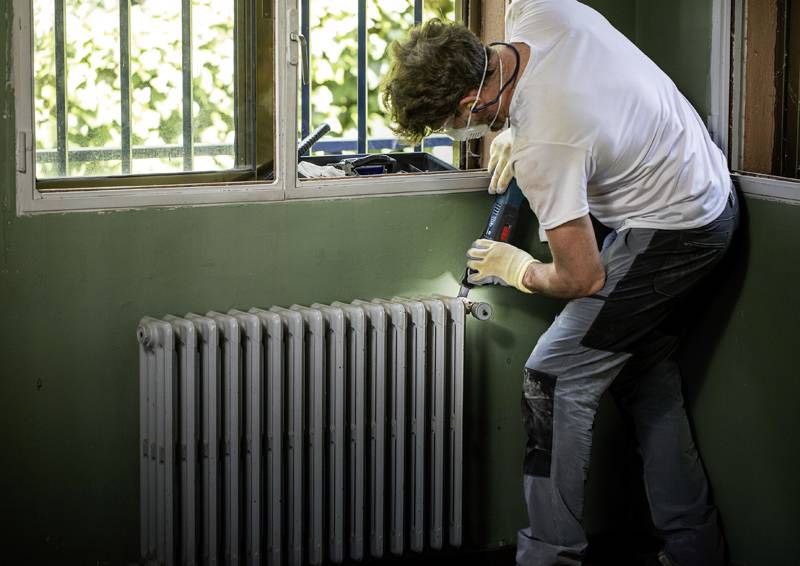 Eine Person mit Handschuhen und einer Maske benutzt einen elektrischen Bohrer, um an einem Heizkörper in einem Zimmer mit grünen Wänden und einem Fenster im Hintergrund zu arbeiten.