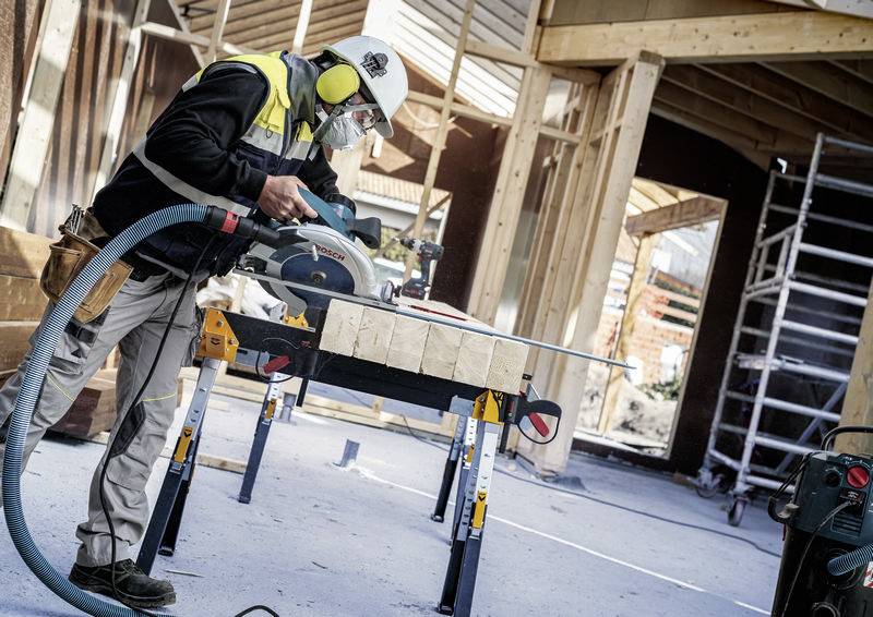 Ein Bauarbeiter in Schutzausrüstung schneidet mit einer Kreissäge Holzbalken auf einer Baustelle zurecht, umgeben von Holzgerüsten.