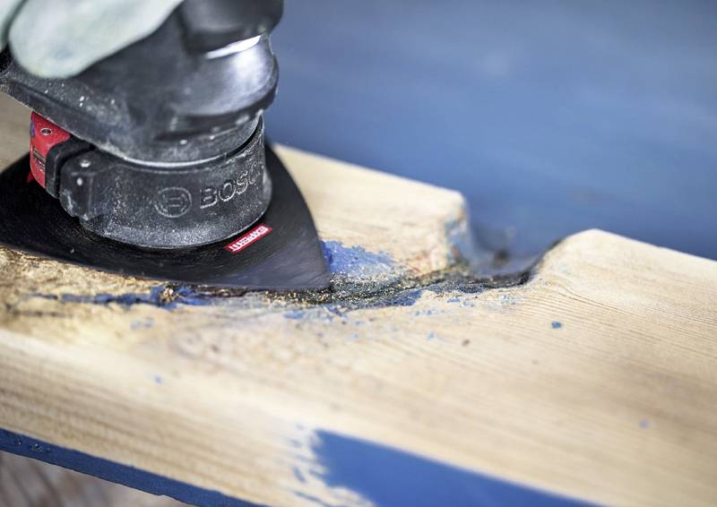 Ein Elektroschleifergerät schleift eine Holzoberfläche ab und entfernt dabei blaue Farbe. Das Gerät ist abgewinkelt, um raue Kanten am Holz zu glätten.