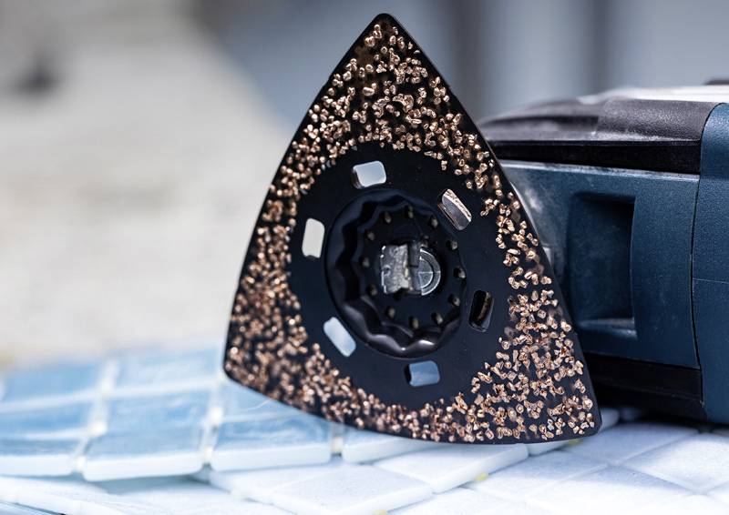Eine Nahaufnahme einer dreieckigen Multitool-Aufsatz mit einer strukturierten Oberfläche, der auf Fliesenmaterial ruht und seine Verwendung zum Schleifen oder Mahlen andeutet.