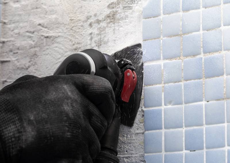 Eine Person mit einem schwarzen Handschuh verwendet ein Elektrowerkzeug, um weißen Fliesenkleber neben einer Wand mit blauen quadratischen Fliesen durchzutrennen.