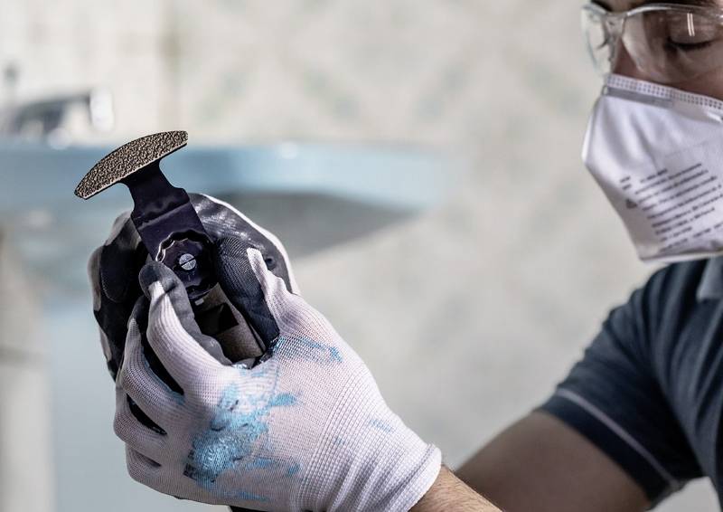 Eine Person trägt Handschuhe und eine Maske und hält ein tragbares Schleifwerkzeug, das mit blauer Farbe bedeckt ist, was auf Do-it-yourself- oder Renovierungsarbeiten hinweist.