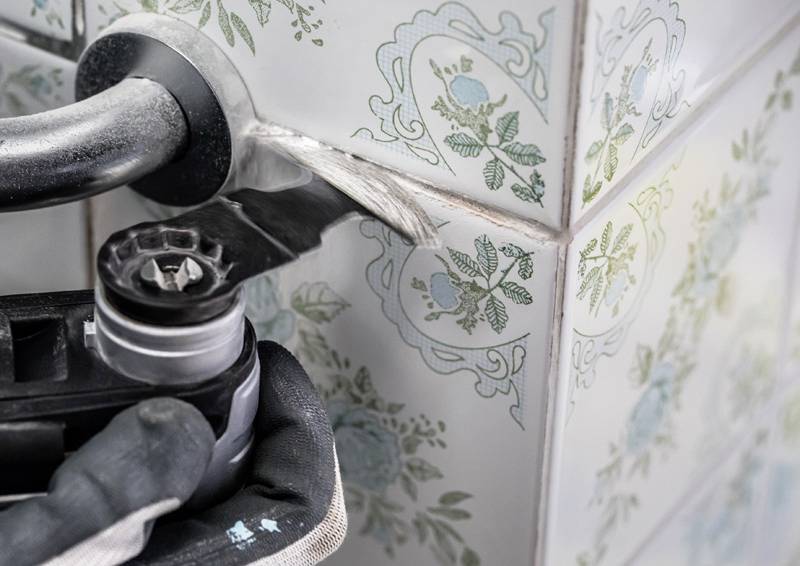 Eine Person mit Handschuhen verwendet einen Winkelschleifer, um Fugenmaterial zwischen dekorativen Blumenkacheln an einer Wand zu entfernen.