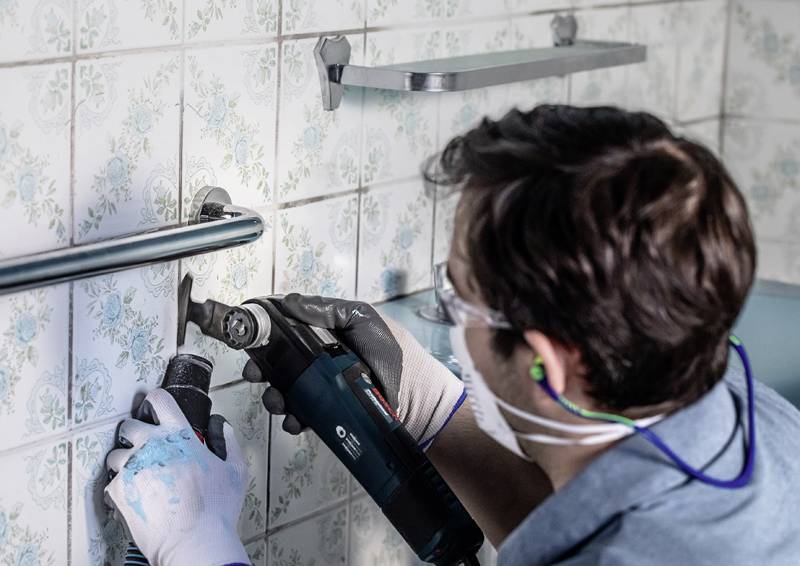 Eine Person in Schutzausrüstung benutzt ein Elektrowerkzeug, um in Badezimmerfliesen zu bohren. Regale und eine Haltestange sind an der Wand sichtbar.
