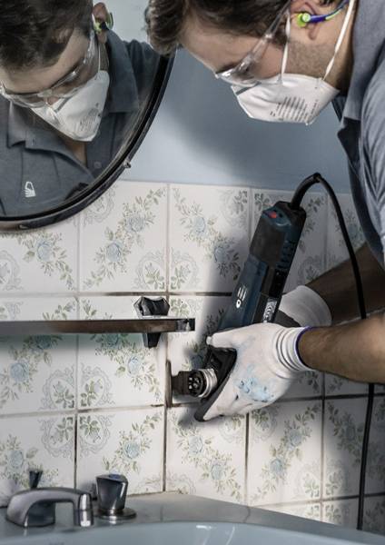 Eine Person in Schutzausrüstung verwendet ein elektrisches Werkzeug zum Schneiden von Fliesen in einem Badezimmer in der Nähe eines Waschbeckens. Schutzbrille und Maske werden getragen.