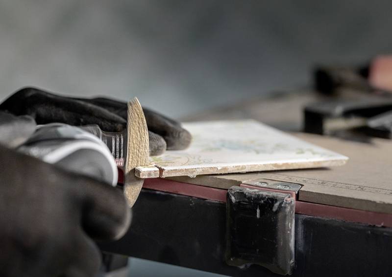 Eine Person nutzt eine Säge, um eine Keramikfliese auf einer Werkbank zu schneiden, dabei trägt sie Schutzhandschuhe. Die Szene deutet auf einen Fokus auf Handwerkskunst hin.