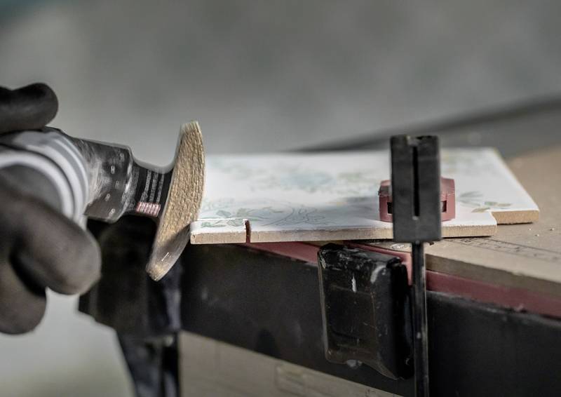 Eine Hand mit einem Handschuh benutzt ein Elektroschleiferwerk mit einer Schneidescheibe, um einen präzisen Schnitt in einer Keramikfliese anzubringen, die in einer Spannvorrichtung befestigt ist.