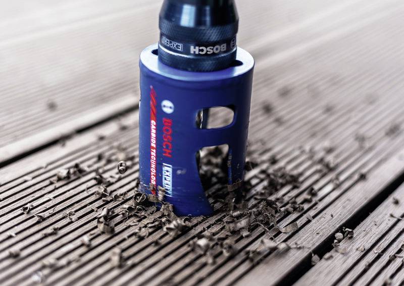 Ein blauer Bohraufsatz mit Rotationsbewegung dringt in Holzdielen ein und erzeugt dabei kreisförmige Holzspäne. Der Markenname „Bosch