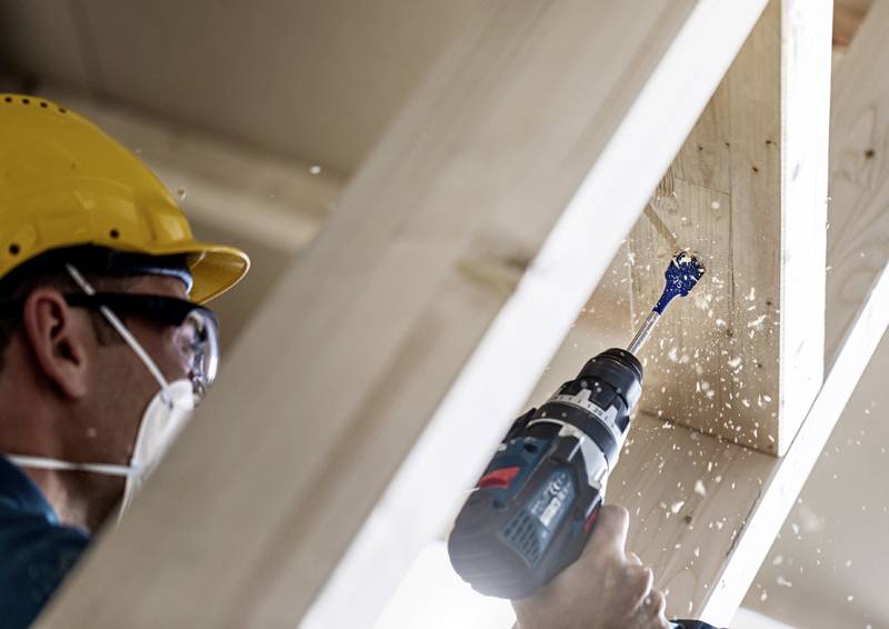 Ein Arbeiter, der Schutzausrüstung trägt, verwendet eine Elektrobohrmaschine an einem Holzbalken, wobei Holzspäne durch die Bohrbearbeitung umherfliegen.