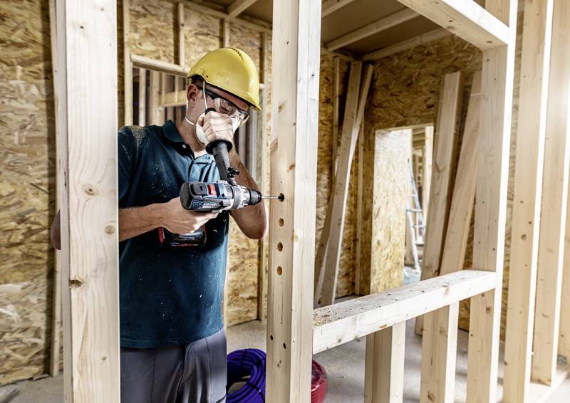Ein Bauarbeiter mit gelbem Schutzhelm und Maske benutzt eine Elektrobohrmaschine an einem Holzrahmen in einem Gebäude im Bau.