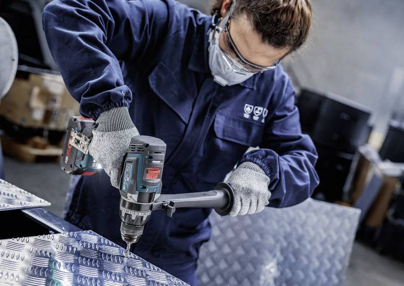 Eine Person in Schutzausrüstung verwendet einen Elektroschrauber an einem Metallblech in einer Industrieumgebung.