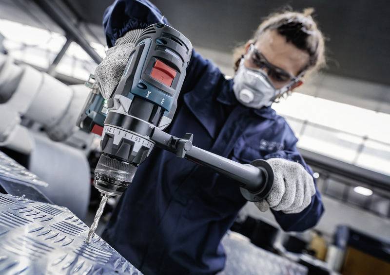 Ein Arbeiter in Schutzausrüstung, der eine Elektrobohrmaschine auf einer Metallfläche in einer Industrieumgebung verwendet.