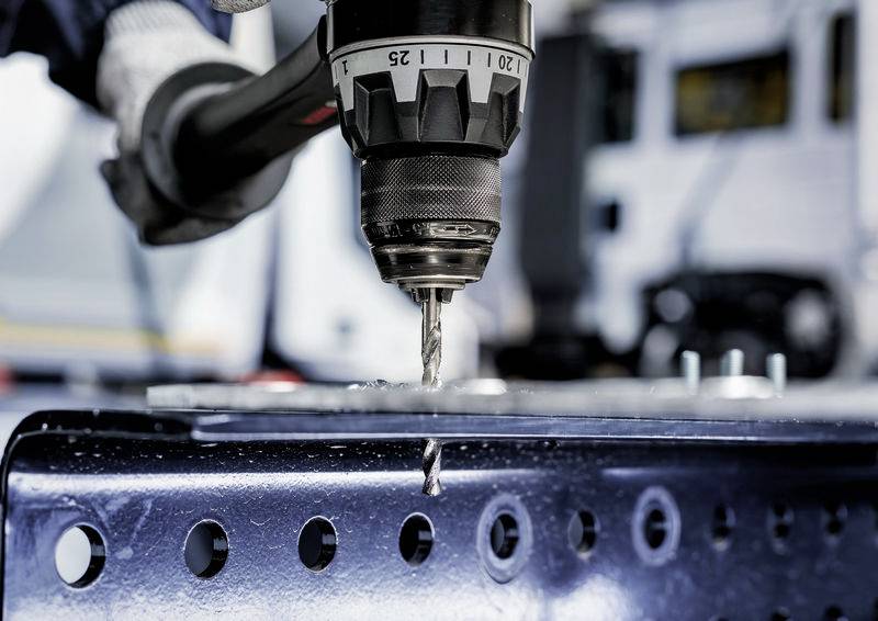 Ein Bohrer mit einem Metallbit macht ein Loch in einem Stahlträger, wobei unscharfe Maschinen im Hintergrund zu sehen sind und industrielle Arbeit demonstriert wird.