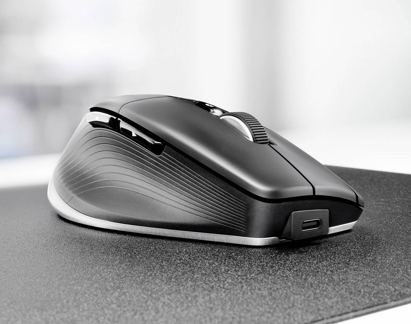 Eine elegante, ergonomische schwarze Computermaus mit strukturiertem Seitengr​iff ruht auf einer grauen Mausmatte, wobei ein USB-Ladeanschluss sichtbar ist.