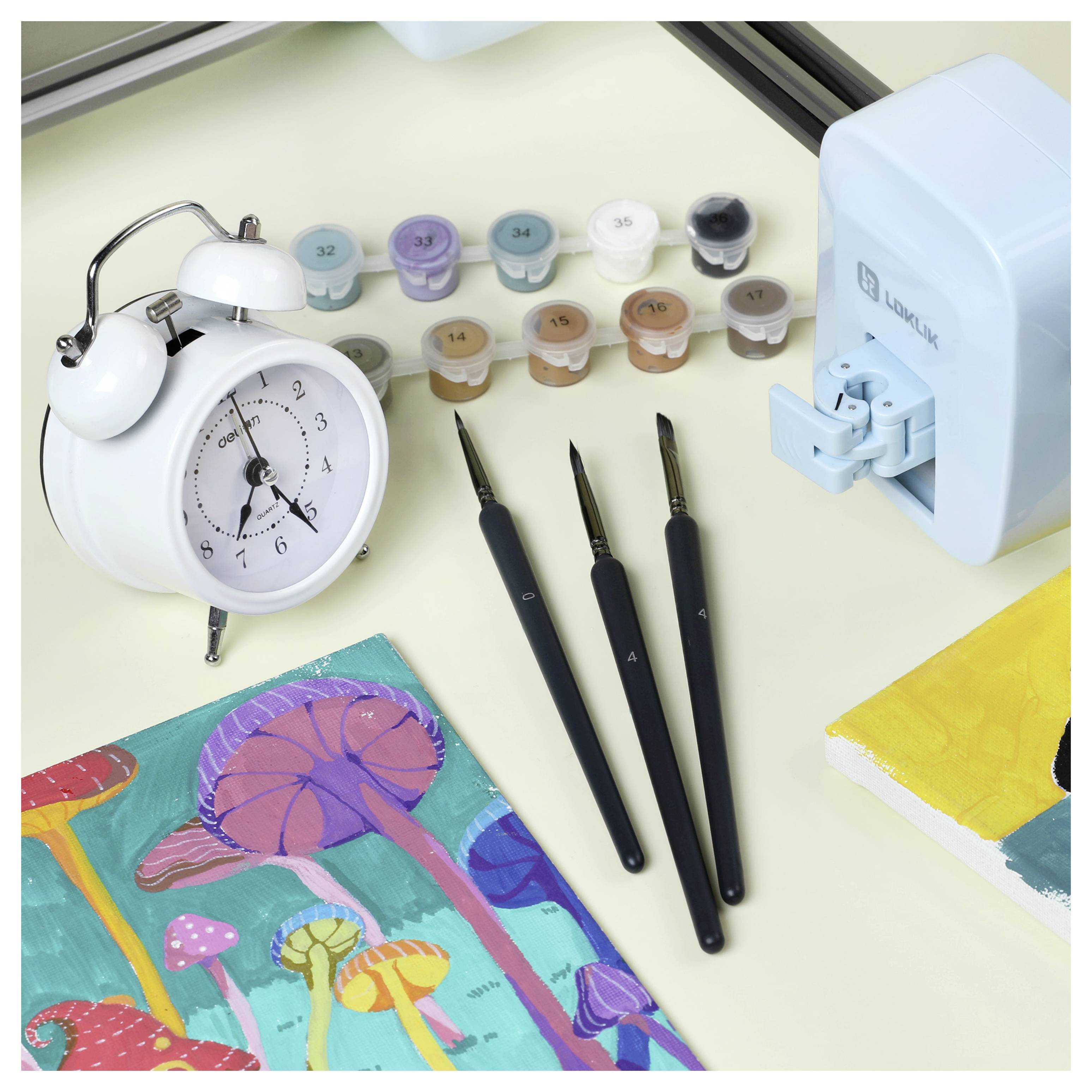 Kunstmaterialien auf einem Schreibtisch umfassen Pinsel, einen Wecker, kleine Farbdosen und ein farbenfohes Gemälde von Pilzen.