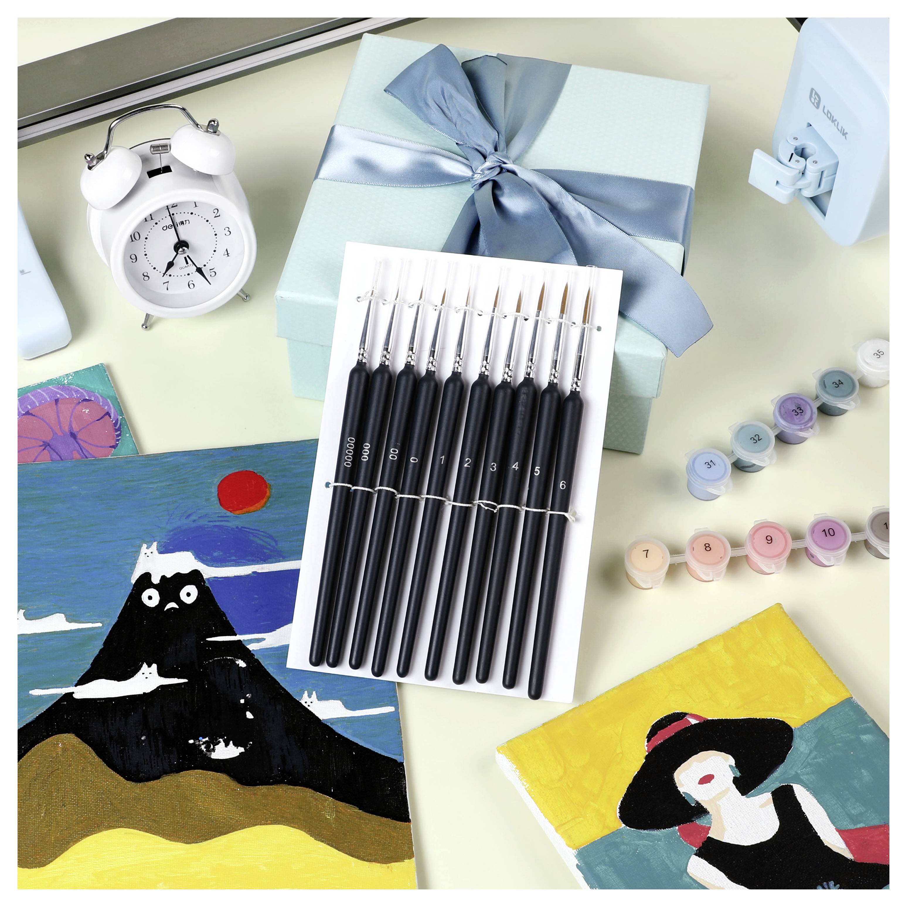 Ein Satz feiner Haarpinsel auf einem Tisch mit verschiedenen Kunstmaterialien, darunter zwei Gemälde, Farbtöpfe, eine Geschenkbox und eine Uhr.