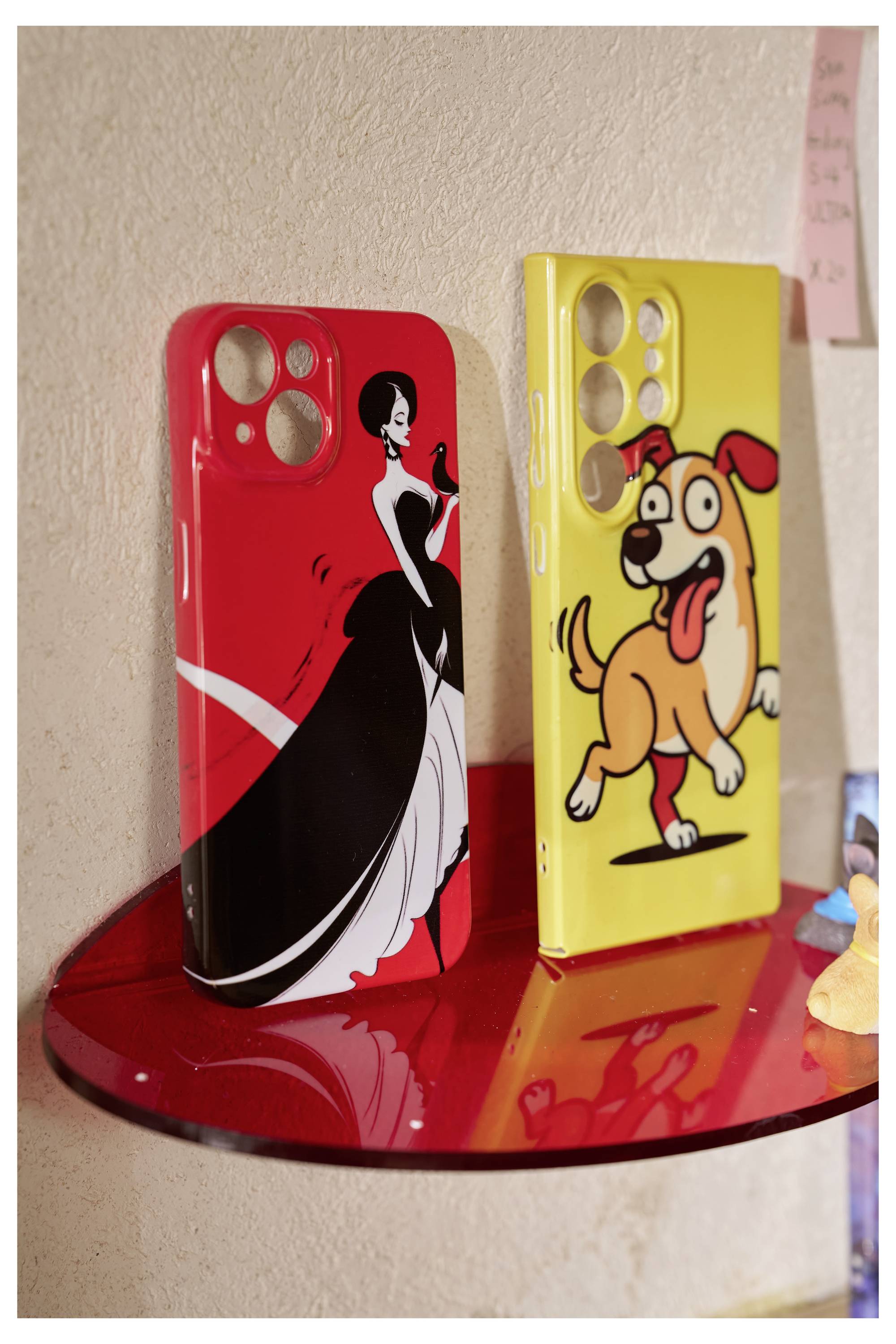 Zwei Handyhüllen auf einem roten Regal: eine rote mit einer Schwarzweiß-Figur in einem Kleid, die andere gelb mit einem Cartoon-Hund.