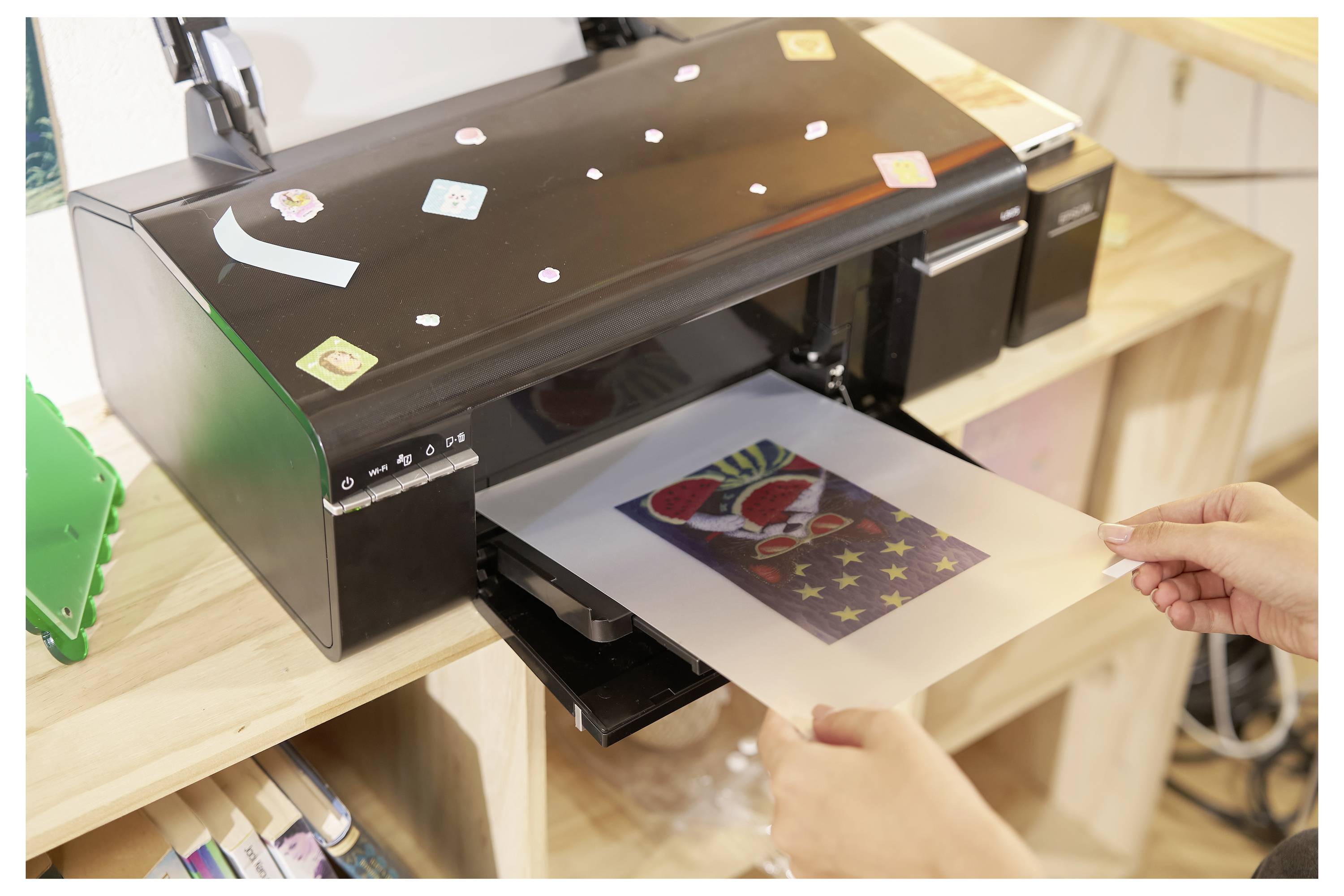 Eine Person entnimmt ein bedrucktes Blatt Papier aus einem Tintenstrahldrucker. Der Druck zeigt eine farbenfrohe Cartoon-Figur mit sternenklarem Hintergrund.