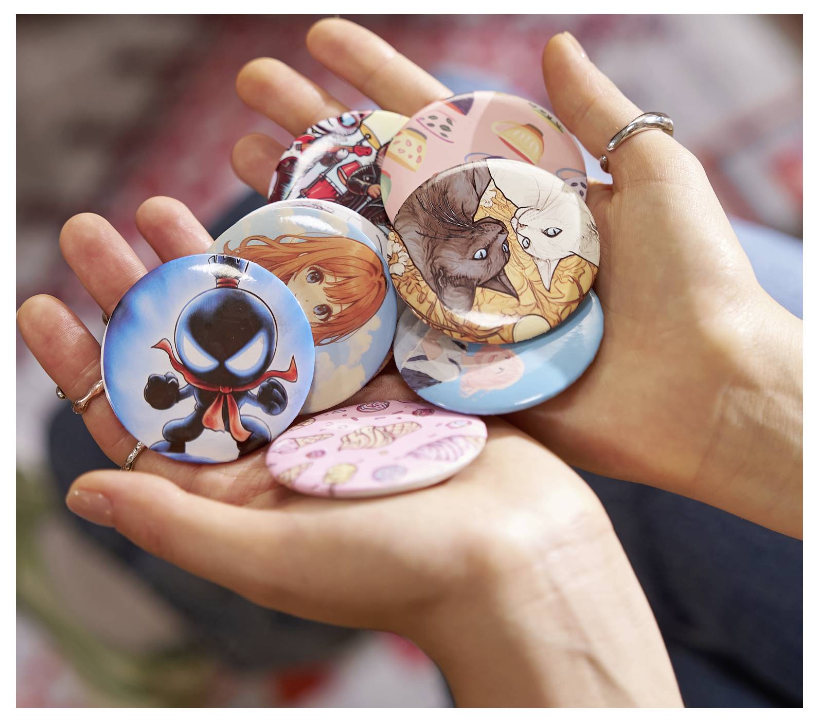 Hände halten eine Sammlung bunter Nadeln mit verschiedenen Cartoon-Designs, darunter abstrakte Charaktere und Zeichnungen von Katzen.